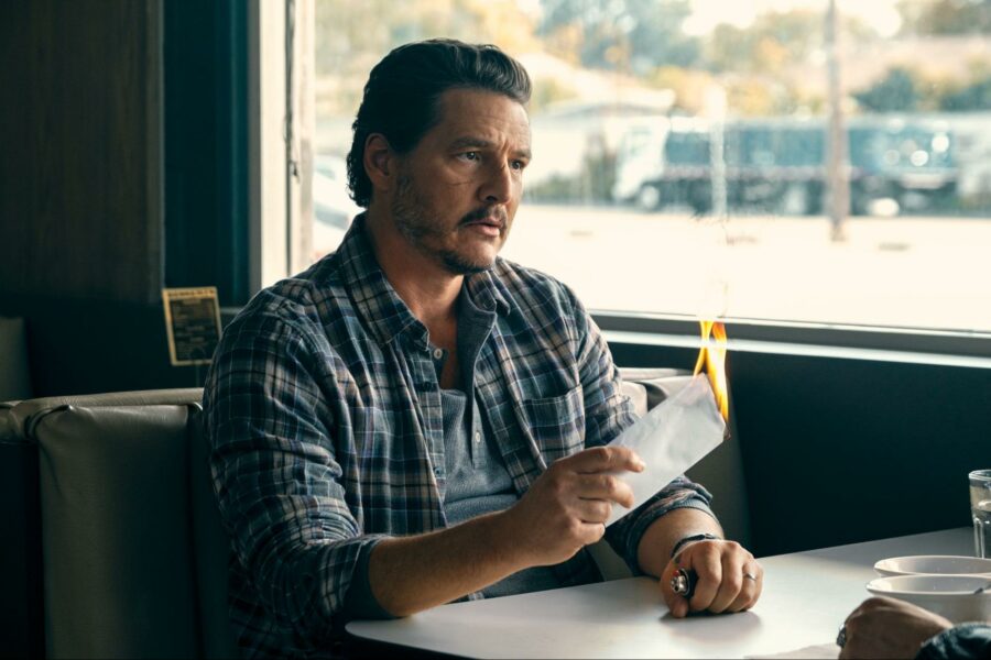 A film still of a man in his 30s to 50s, sitting at a diner table and holding a white envelope which is on fire