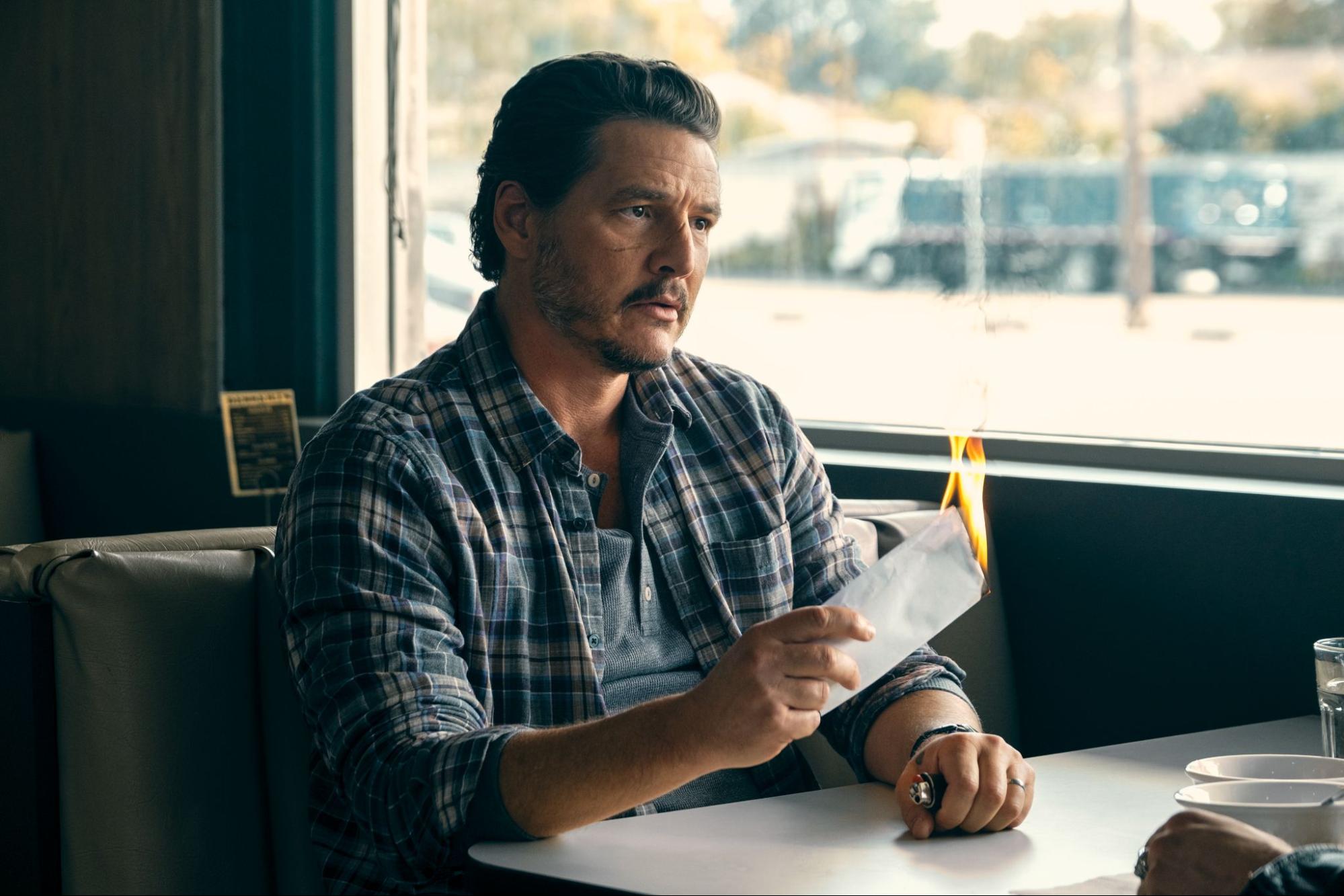 A film still of a man in his 30s to 50s, sitting at a diner table and holding a white envelope which is on fire
