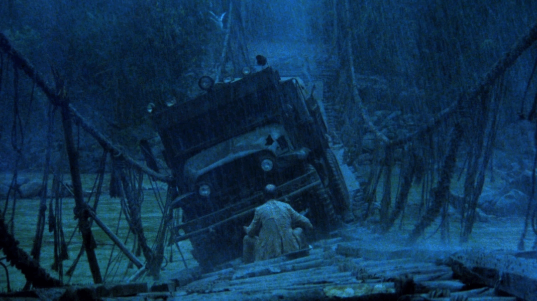 A film still of a very dark scene tinted in blue. A rickety bridge is collapsing under the weight of a truck and there is a man trying to hold the truck up